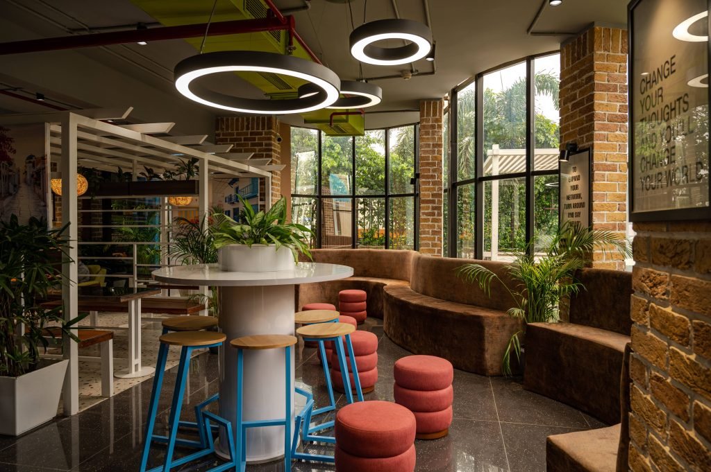 Office interior featuring numerous plants, sustainable decor elements, cozy seating, and large windows promoting natural lighting.