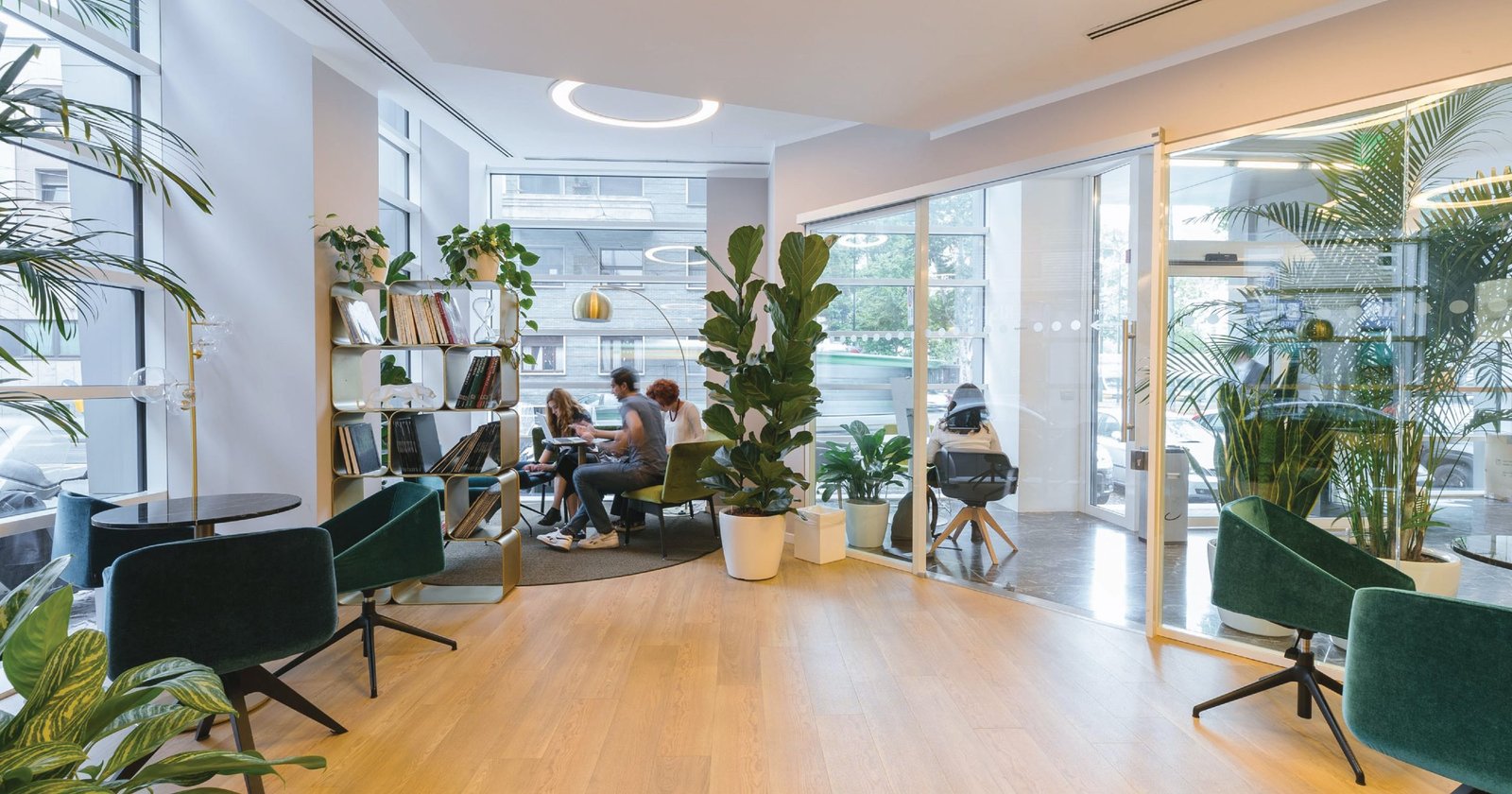 A bright, spacious collaborative office workspace featuring comfortable seating, large windows, abundant greenery, and open-plan design.